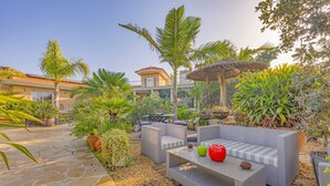 Eine Terrasse im Freien mit Steinböden, einem grauen Sofa, einem Tisch mit Dekoration und einem Reetdachschirm.