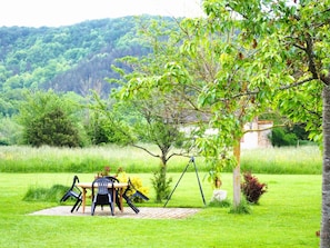 Ein Picknicktisch mit Stühlen und einer Schaukel in einem grasbewachsen Feld, im Hintergrund Bäume und eine Hügelkette.