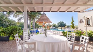 A covered outdoor area with a round table and chairs, a thatched umbrella, and a pool in the background. - Villa 'Apollonion' with Private Pool, Wi-Fi and Air Conditioning (Siracusa)