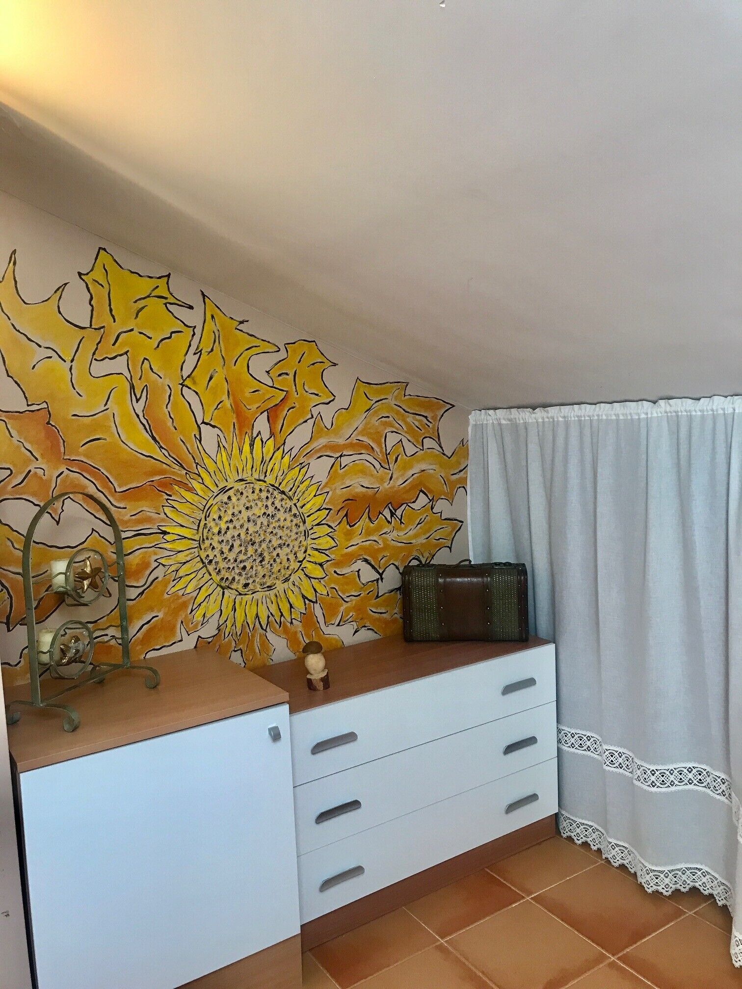 Una habitación con un aparador blanco, un mural grande con girasoles, un objeto decorativo pequeño y una cortina blanca.