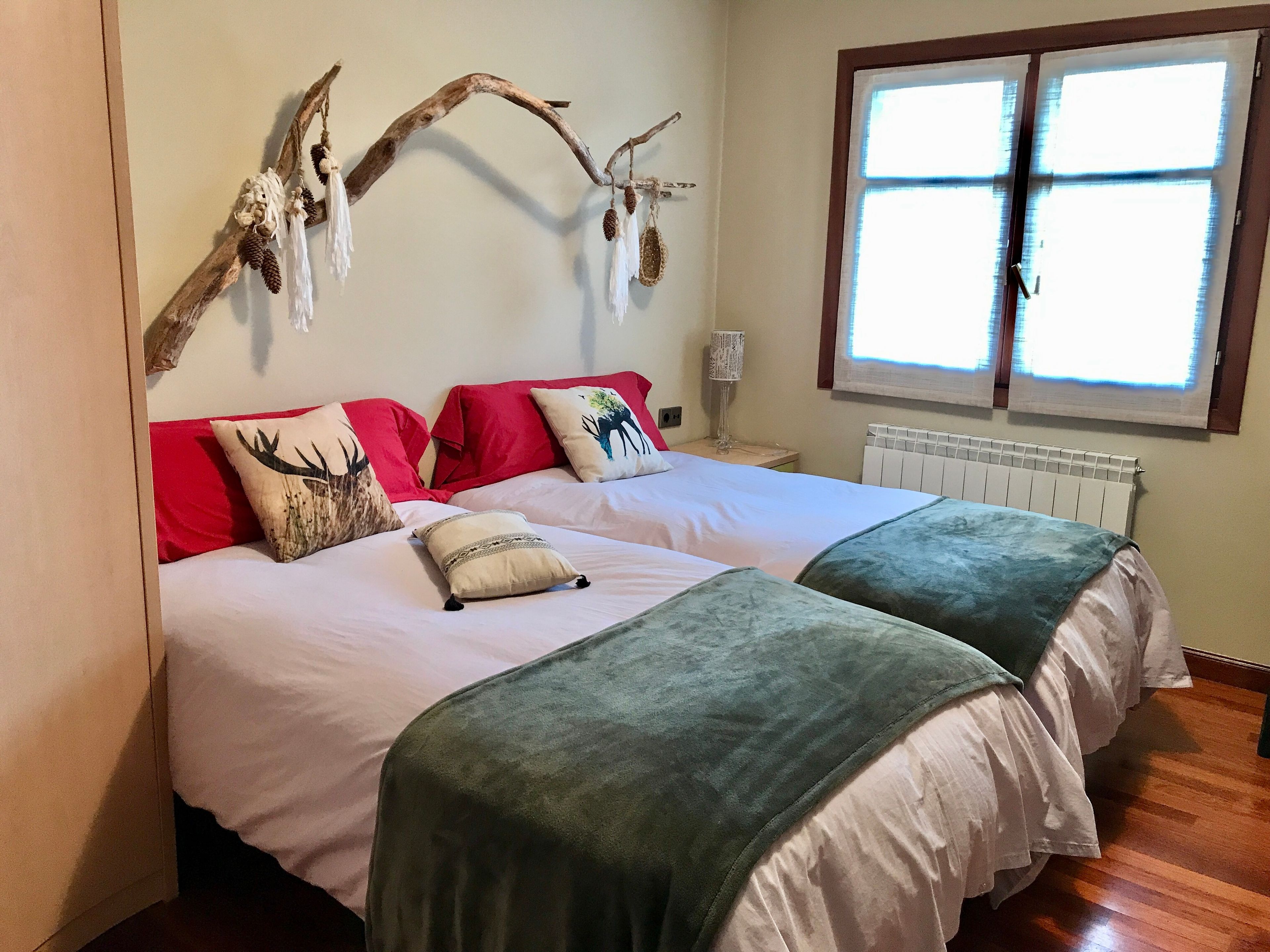 A double bed with white bedding and green throw pillows, a decorative wall piece above the bed, and a window with blinds.