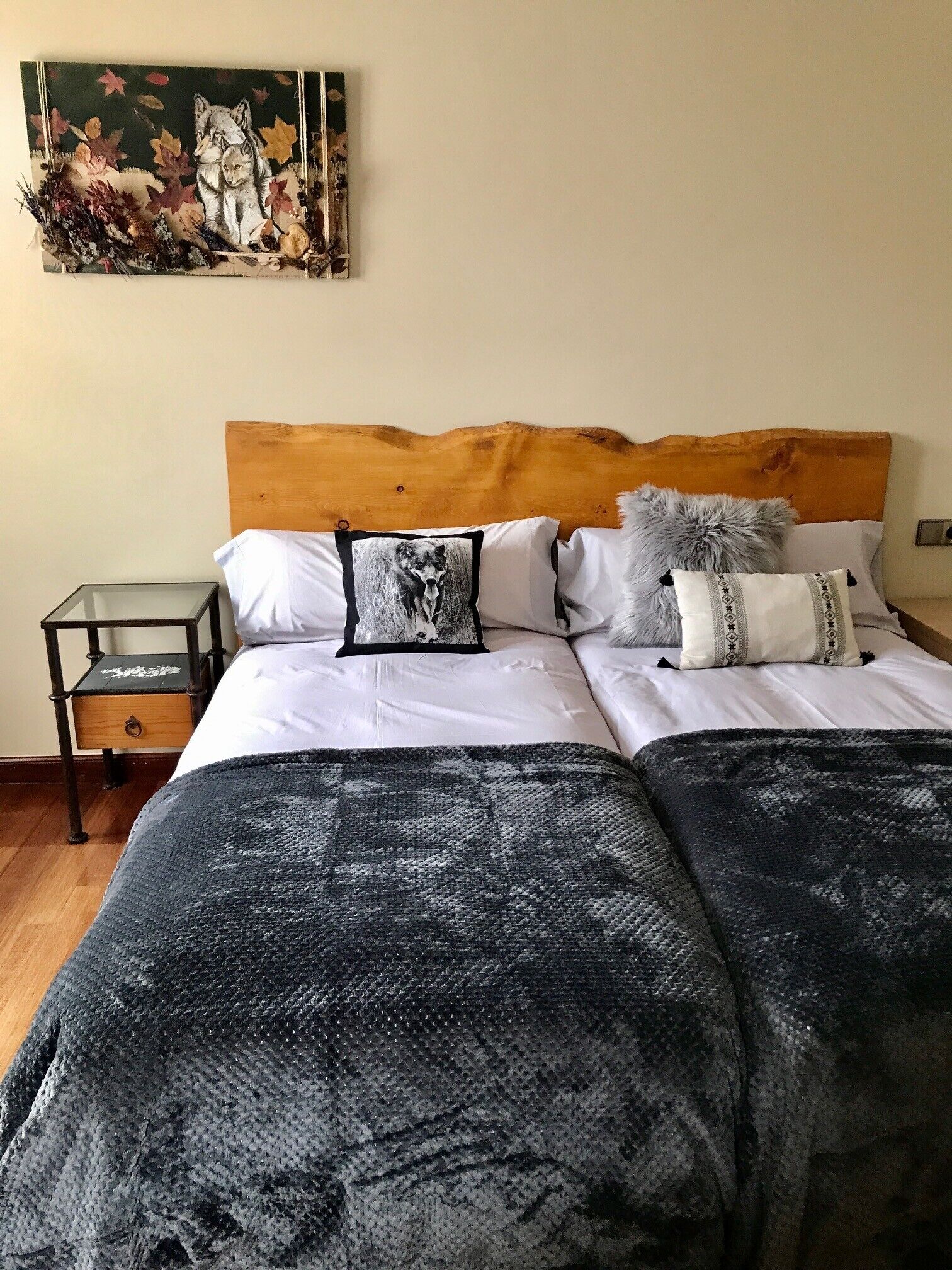 Un dormitorio con cabecera de madera, cama con ropa blanca, varios almohadones y una mesita de vidrio, además de un cuadro colgado en la pared.