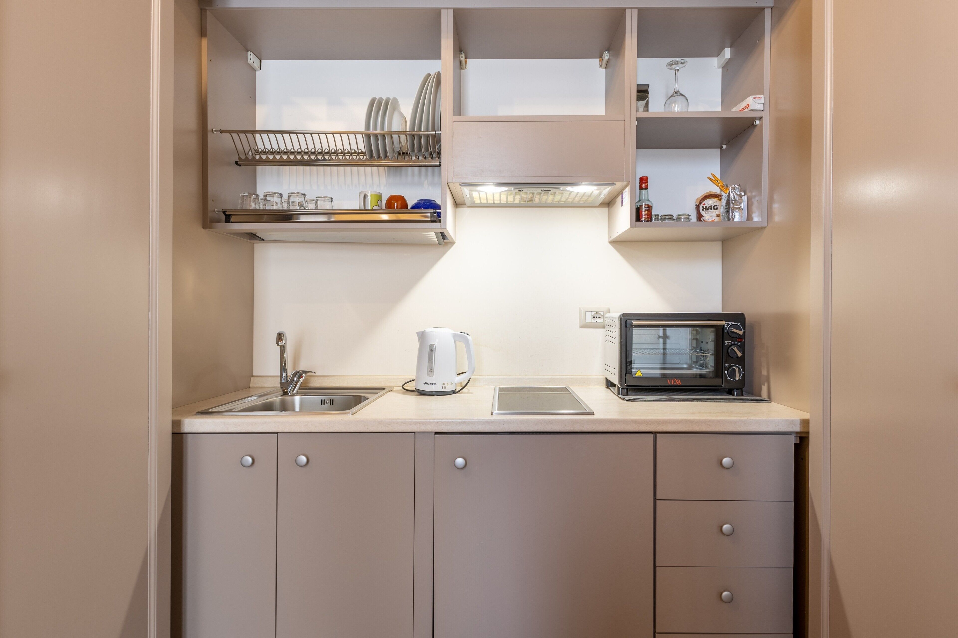 A compact kitchenette with a sink, kettle, microwave, and shelves for dishes and glasses.