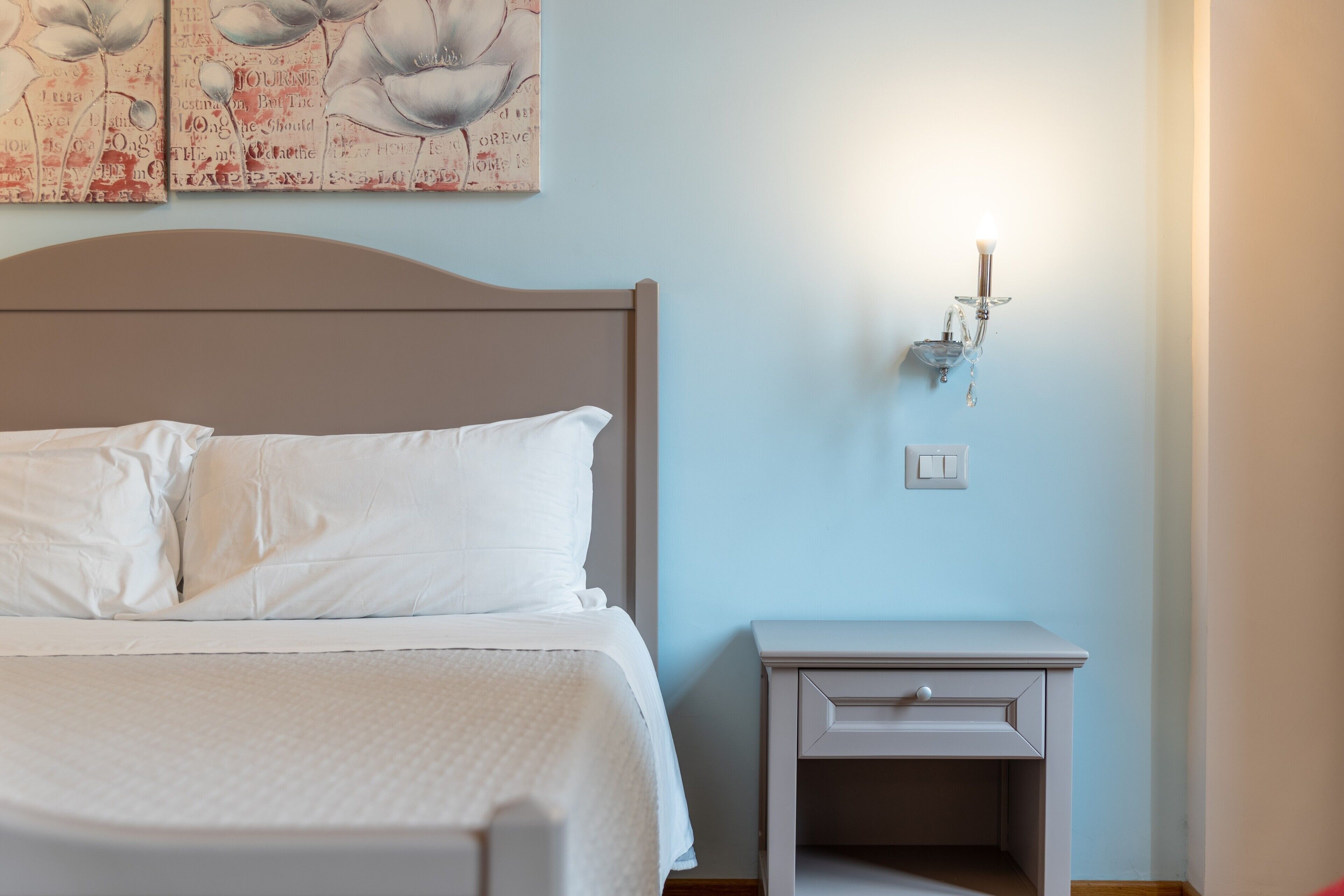 A neatly made bed with white linens, a bedside table with a drawer, and a wall-mounted light fixture.