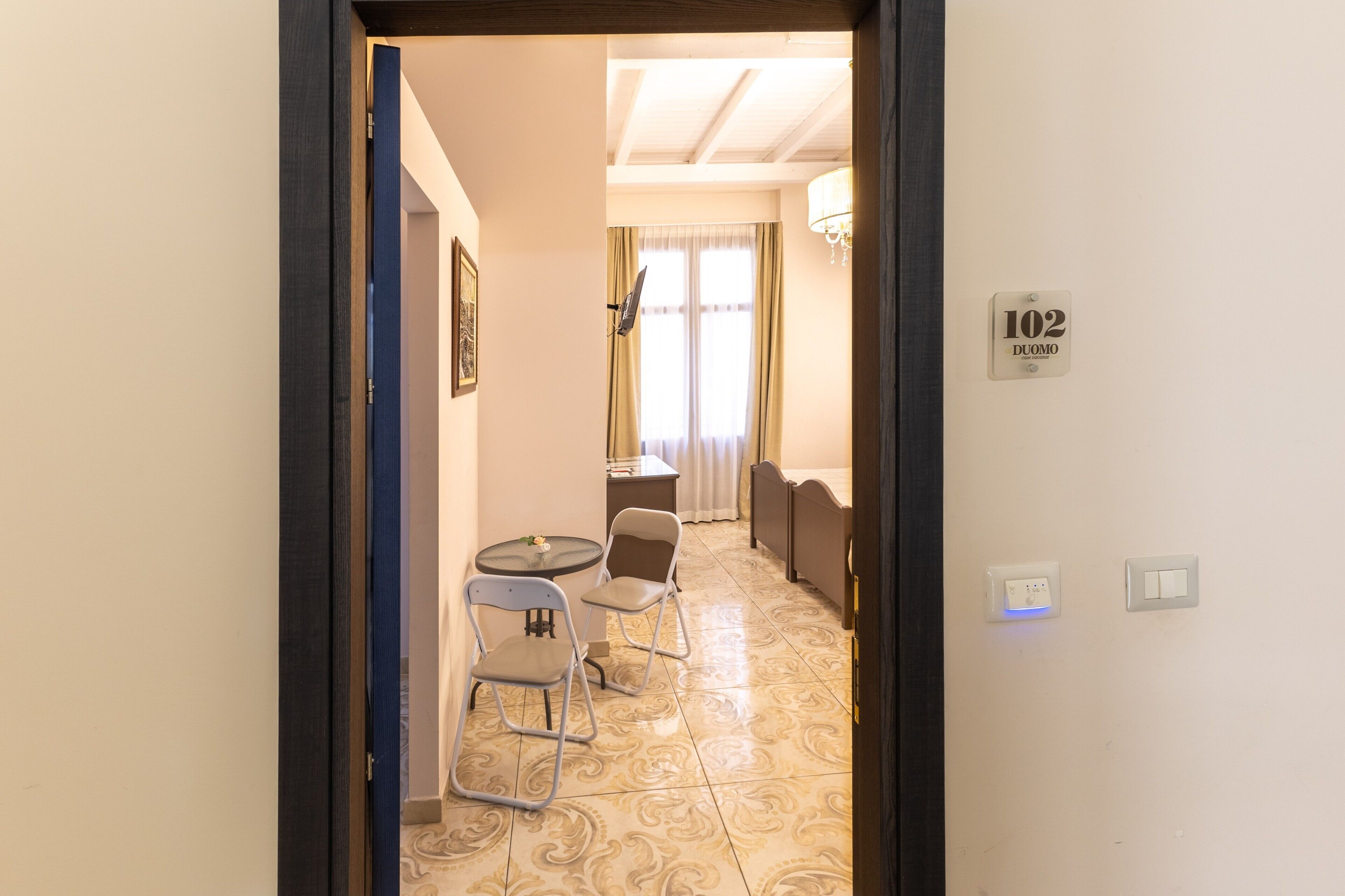 Ein Hotelzimmer mit Bett, einem kleinen Tisch mit Stühlen, einem Fenster mit Vorhängen und einem an der Wand befestigten Lichtschalter.