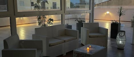 A rooftop terrace with wicker furniture, a table with candles, and large windows overlooking a cityscape.