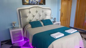 A bedroom with a tufted headboard, a bed with teal and white bedding, a nightstand with a lamp, a dresser, and a painting on the wall.