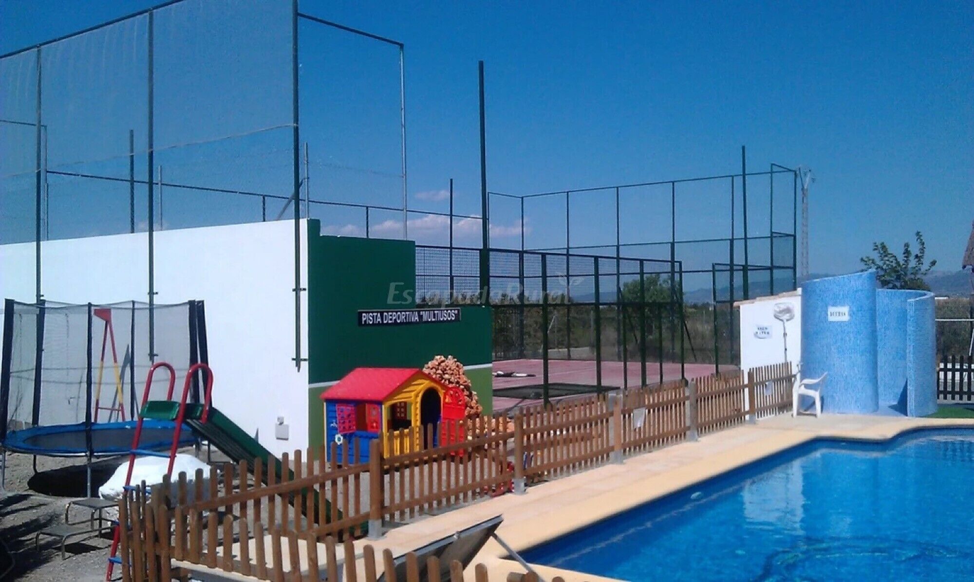 Un área al aire libre cercada con piscina, un pequeño parque infantil y una cancha deportiva.