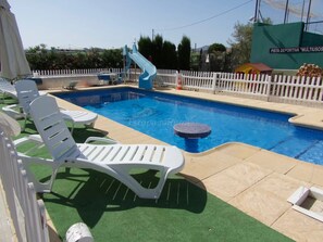 Una piscina con tobogán, sillas de descanso blancas y un área delimitada por una valla.