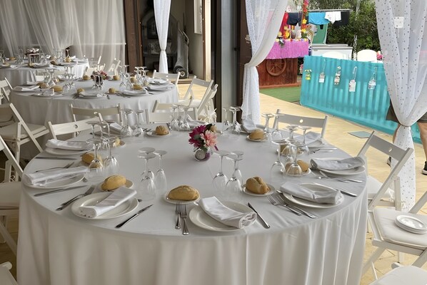 Mesas redondas cubiertas con manteles blancos, sillas blancas y cubiertos.
