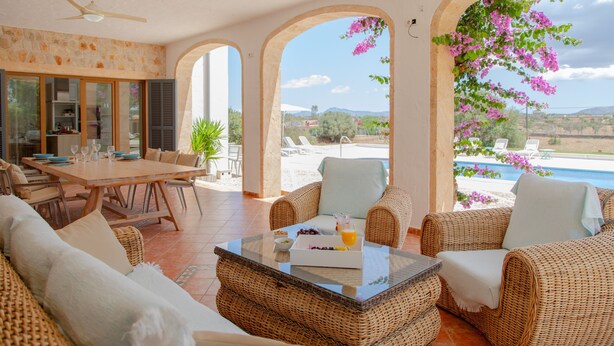 Un vaste espace de vie comprenant une table à manger, des meubles en osier et une vue sur une piscine et un paysage verdoyant.