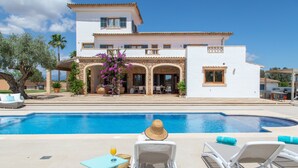 Un espace aménagé au bord de la piscine, avec des chaises longues, une table et une vue sur une grande maison dotée de nombreuses fenêtres et d’un patio couvert.