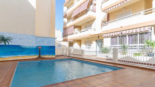 Una piscina con solárium empedrado, un mural con escena de playa y un edificio con balcones y toldos.