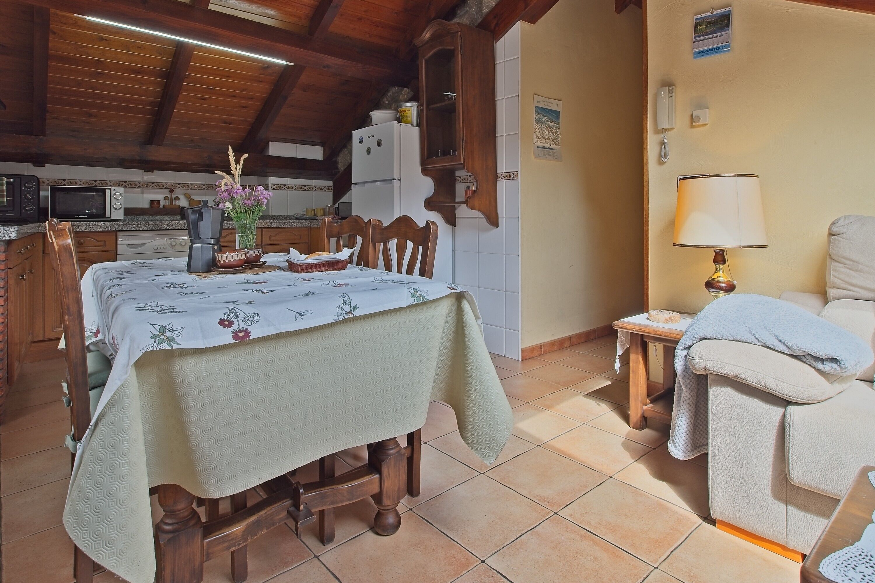 A dining area with a table set for four, a kitchen in the background, and a small bedside table with a lamp.