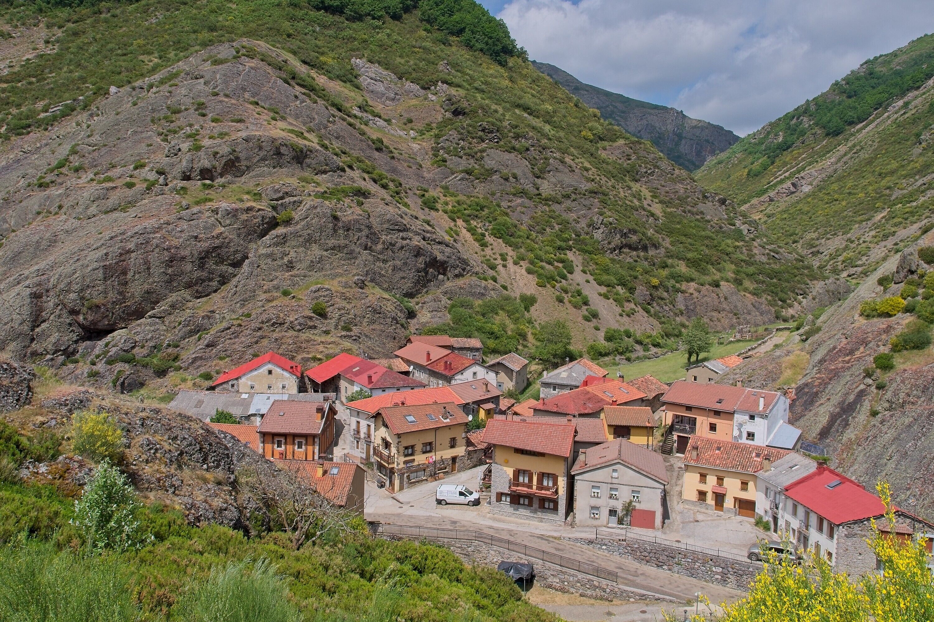 Un village perché sur une colline, avec des bâtiments traditionnels, une route pavée et une voiture blanche garée devant.