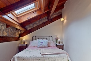 A bedroom with a skylight, stone wall, wooden beams, a bed with floral bedding, and bedside lamps. - Eagle's Nest Country House in Picos de Europa with Mountain Views (Portilla de la Reina)