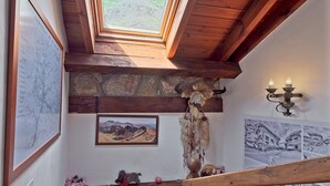 Una habitación con techo de madera, pared de piedra, un mapa, una fotografía enmarcada, un atrapasueños y un perro pequeño.