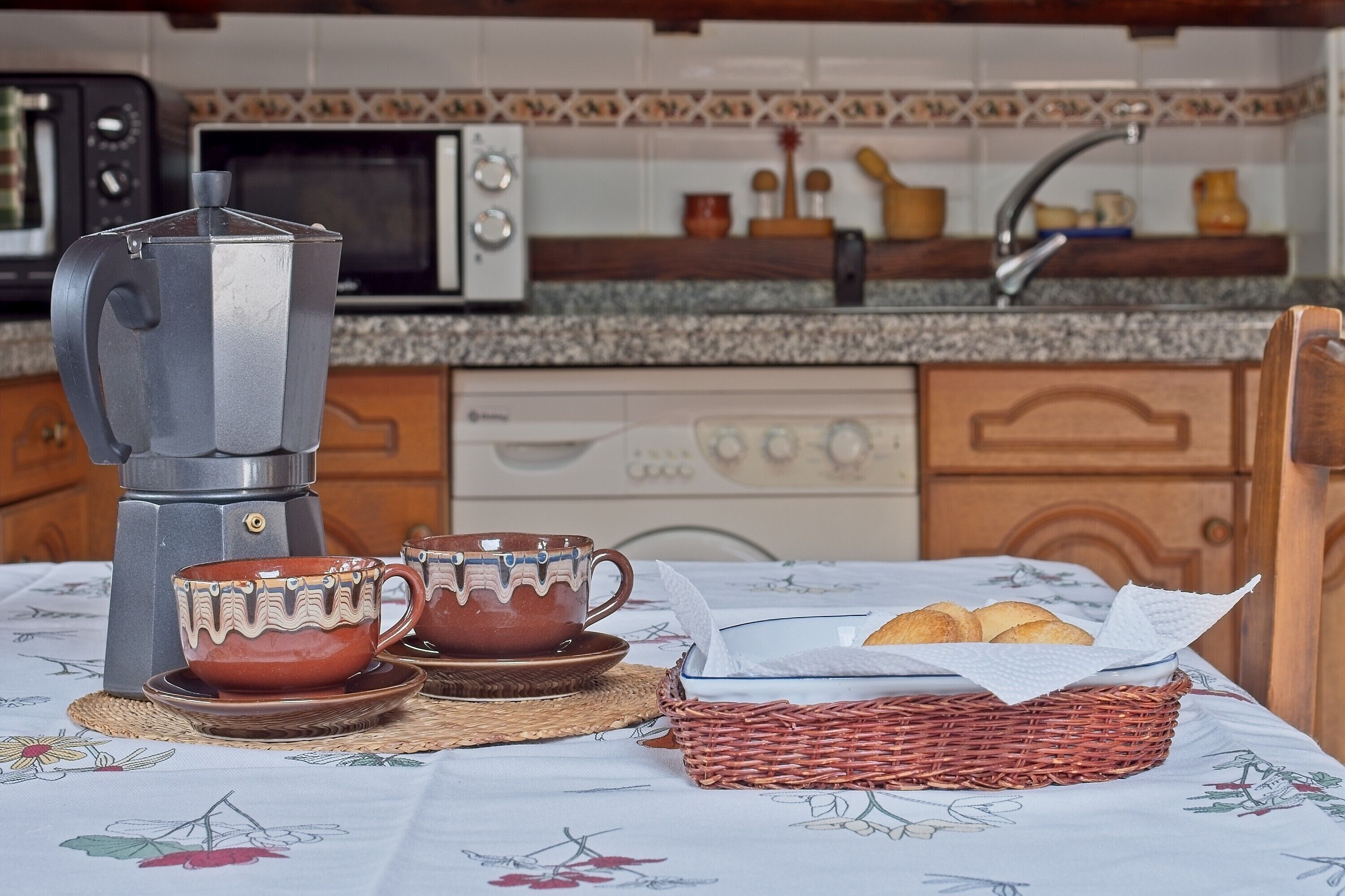 Una cocina con cafetera, dos tazas, una cesta de pan y un microondas.