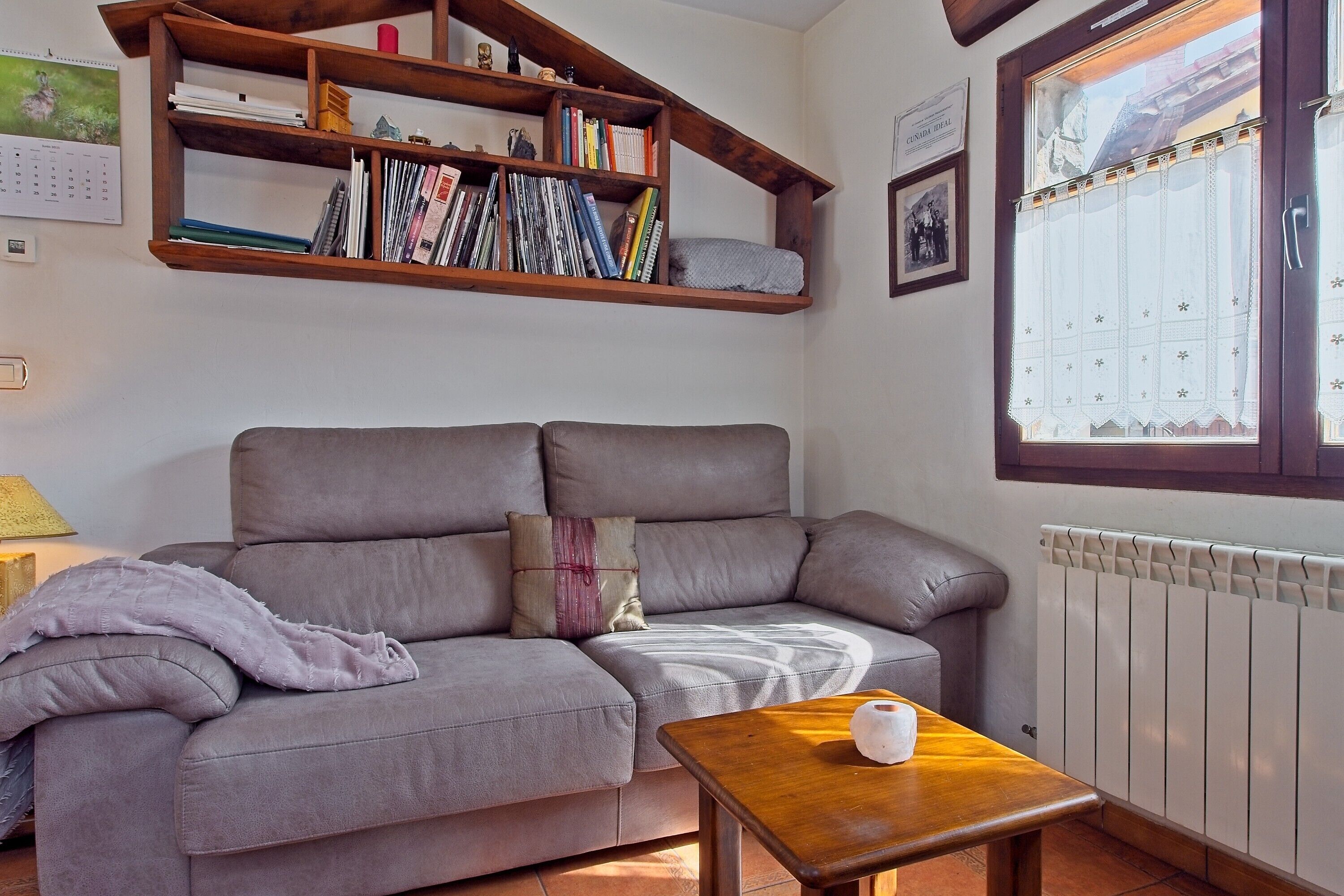 A living room with a grey sofa, a wooden coffee table, a bookshelf, and a window with a curtain.