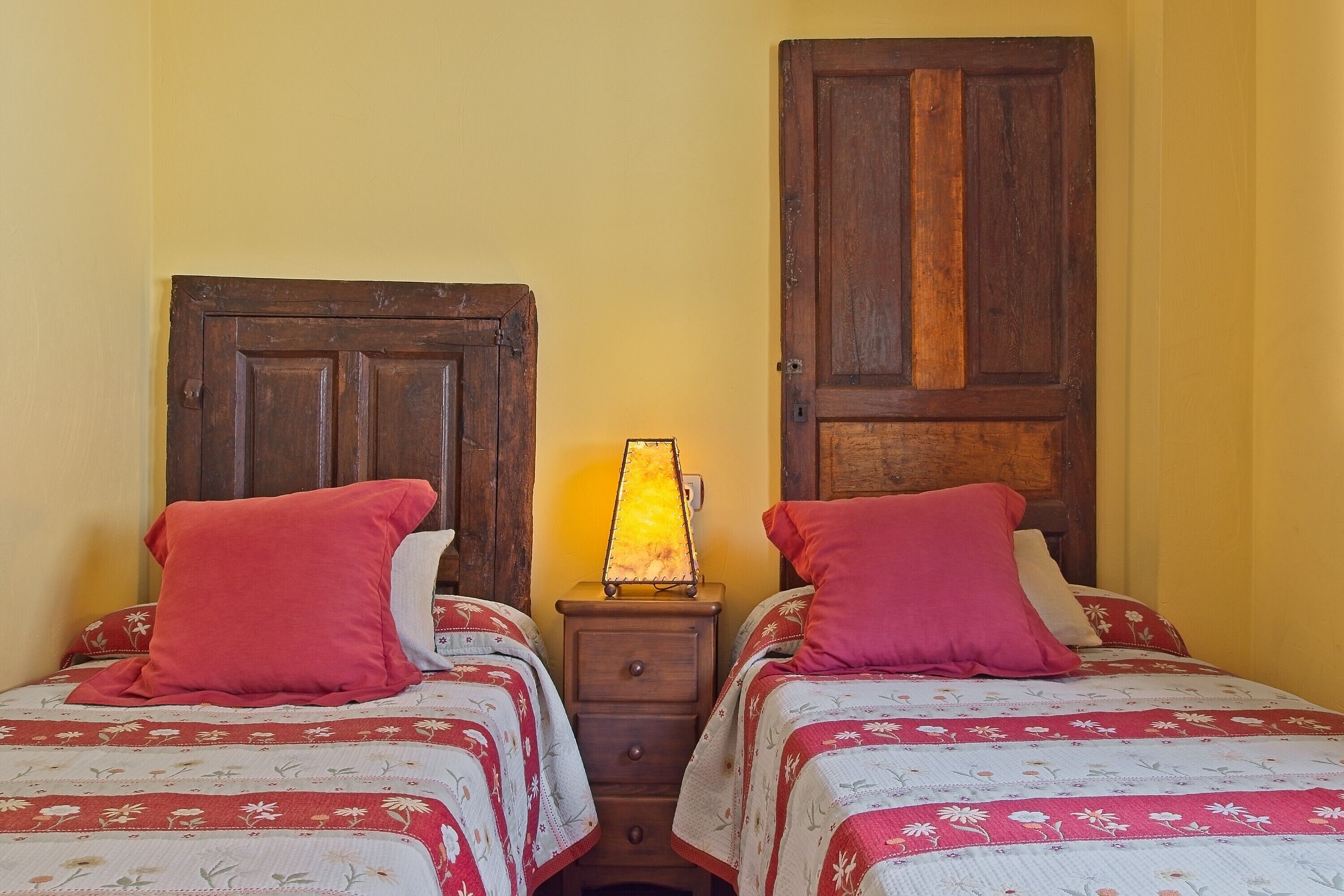Duas camas com roupa de cama floral e almofadas vermelhas, uma mesa de cabeceira de madeira com abajur, e uma porta de madeira grande.