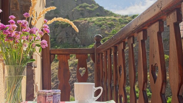 Ein Tisch mit einer weißen Tasse und Untertasse, einer Blumenvase und Gläsern auf einem Balkon mit Blick auf eine gebirgige Landschaft.