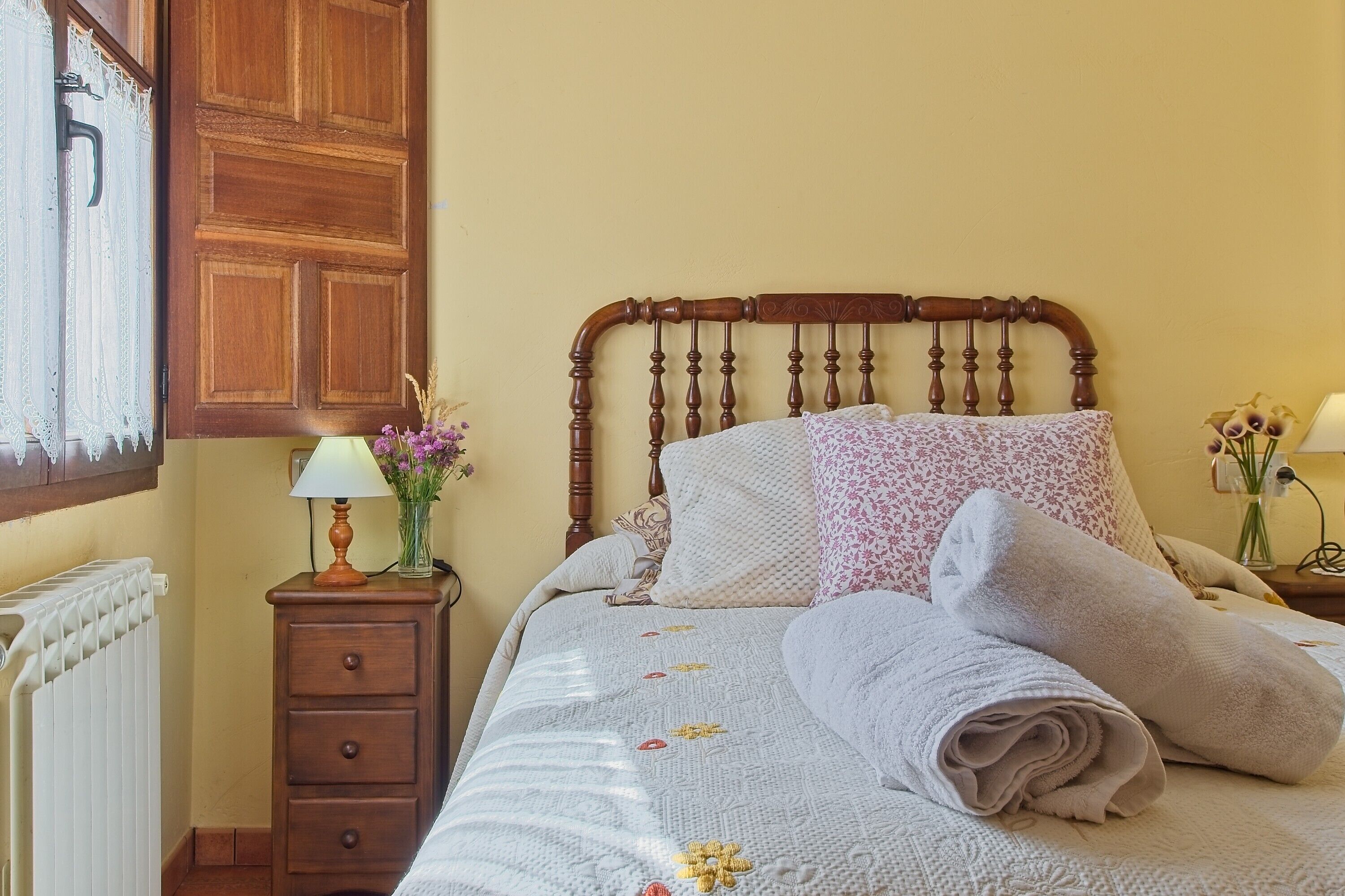 Una cama bien tendida con un cabecero de madera, una mesita de noche con una lámpara y un radiador.