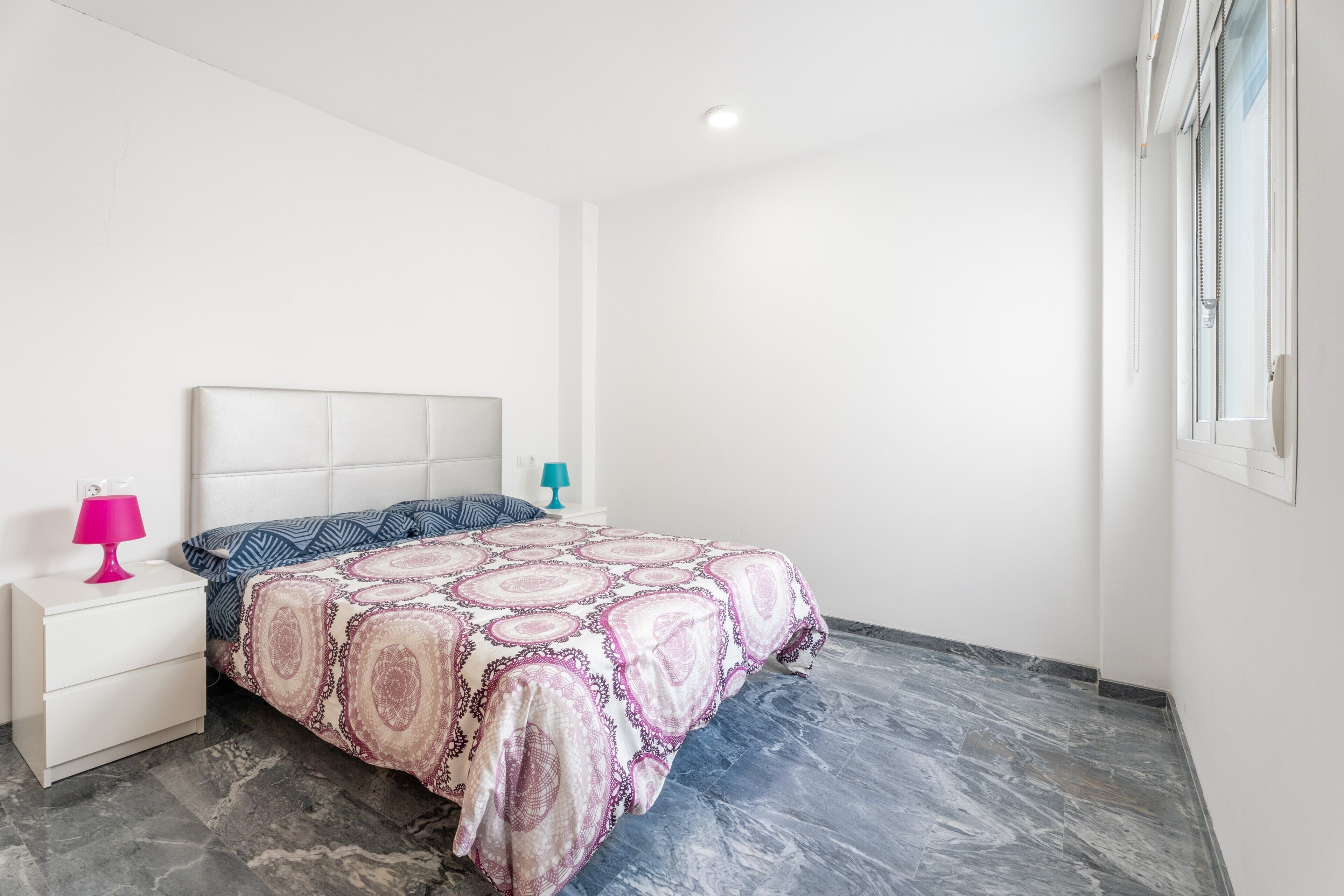 Un dormitorio con cama, mesita de noche, lámpara y ventana con persianas.