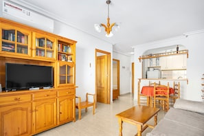 Una sala de estar con televisor, armarios de madera, una mesa de comedor y una zona de cocina.