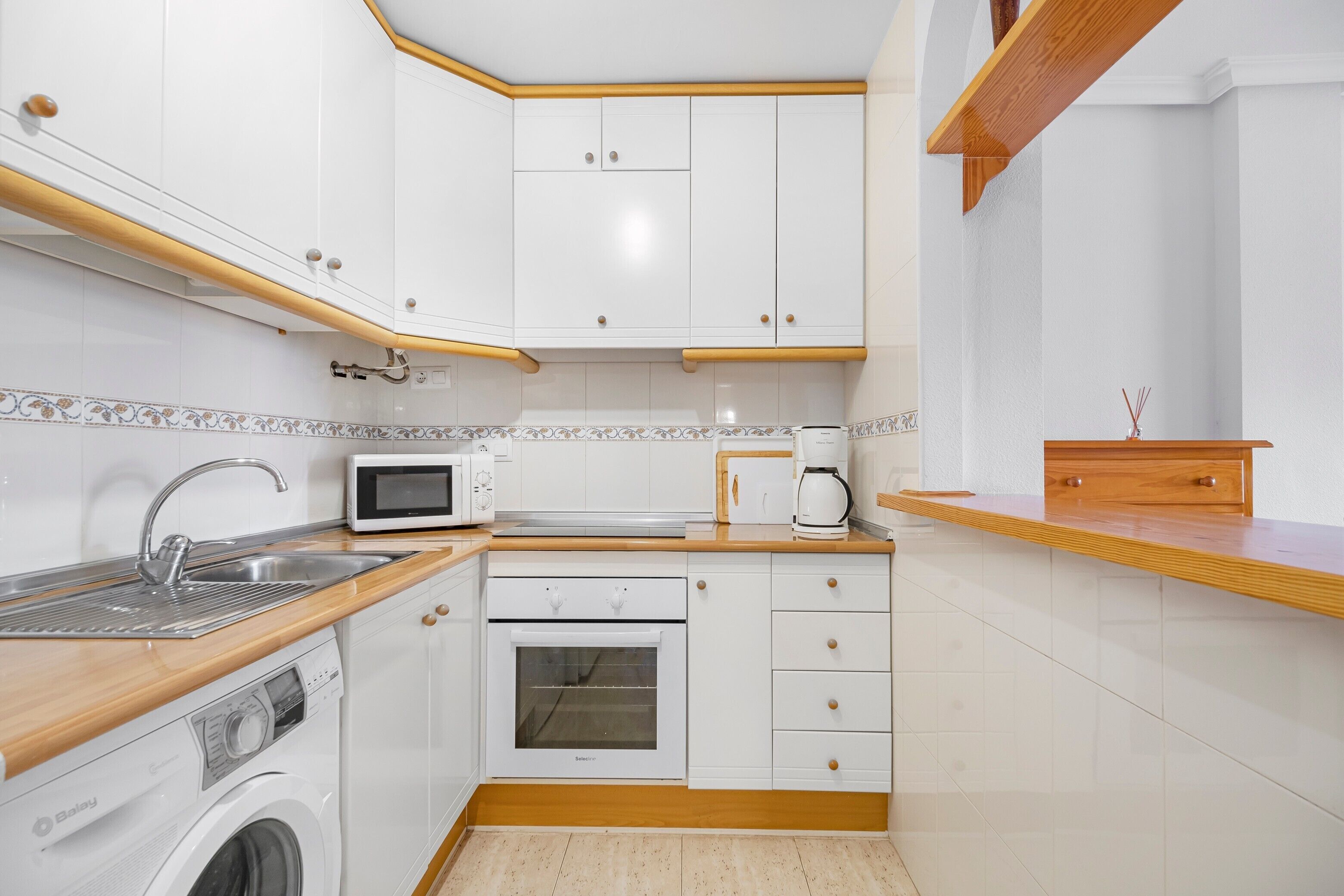 A compact kitchen with white cabinets, a built-in microwave, a coffee maker, and a washing machine.