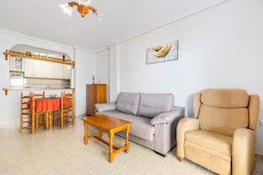 Una sala de estar con sofá, sillón, mesa de comedor de madera y una zona de cocina con armarios y encimera.
