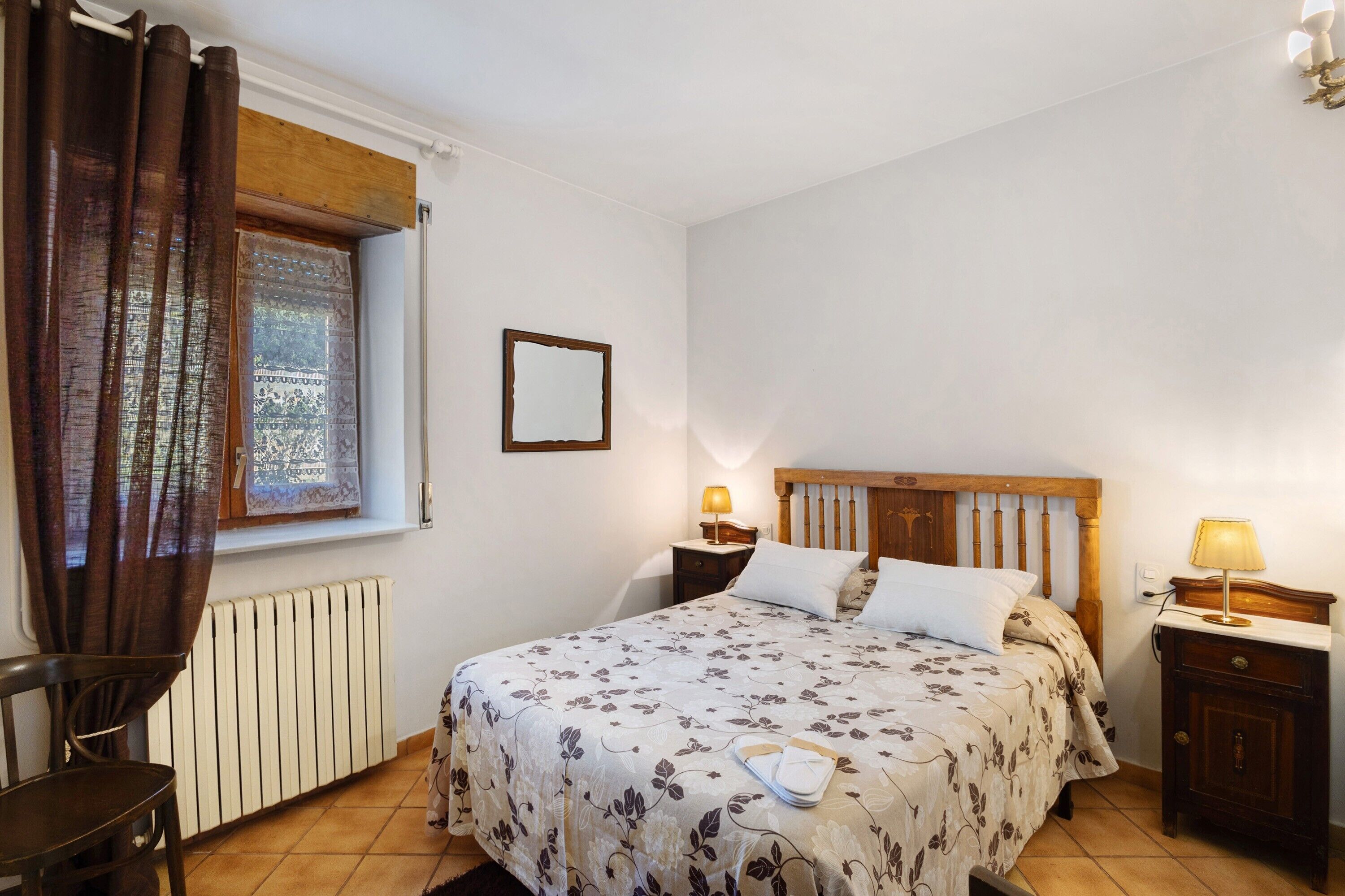 Un dormitorio con cama, ventana con cortinas, mesita de noche con lámpara, radiador y una silla.