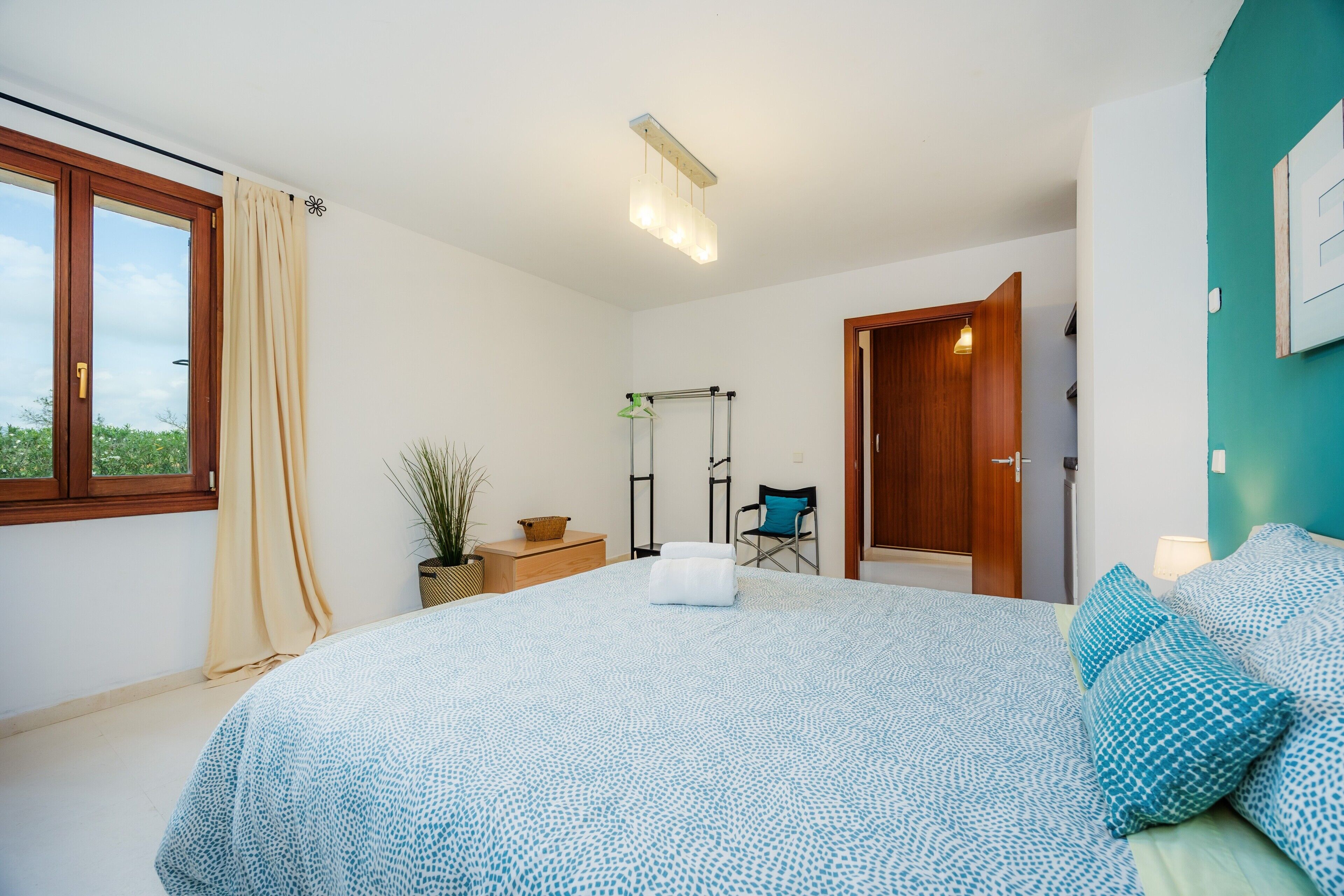 A bedroom with a large bed, a window with curtains, a plant, a chair, and a door.