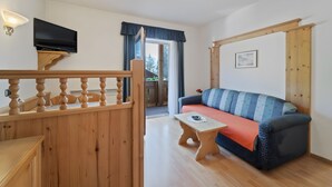 Une pièce avec un escalier en bois, un canapé, une petite table, un téléviseur fixé sur un meuble en bois et une fenêtre avec des rideaux.