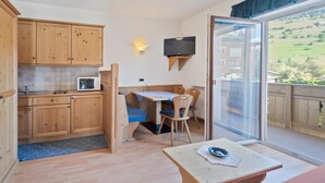 Une pièce dotée de meubles de rangement en bois, d’un four à micro-ondes, d’une table avec des chaises, d’un téléviseur fixé au mur et d’une porte-fenêtre coulissante donnant sur un balcon.