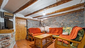 A rustic room with exposed wooden beams, a stone wall, a wooden table, and a sofa with orange cushions.