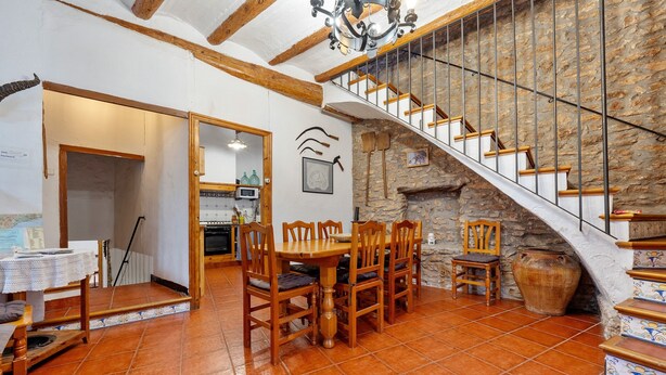 A dining area with a wooden table and chairs, a kitchen in the background, and a stone staircase.