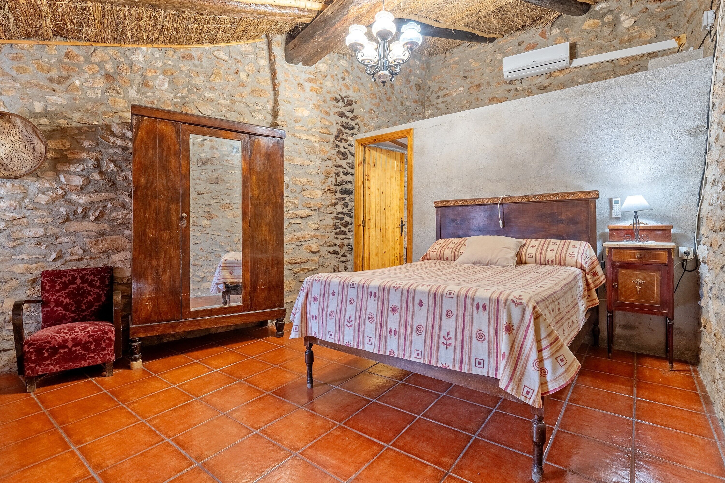 Un dormitorio con pared de piedra, cama de madera, una silla roja, armario de madera y suelo de baldosas.