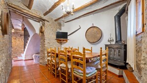 A rustic dining area with a stone wall, wooden chairs, a television on a stand, and a wood-burning stove.