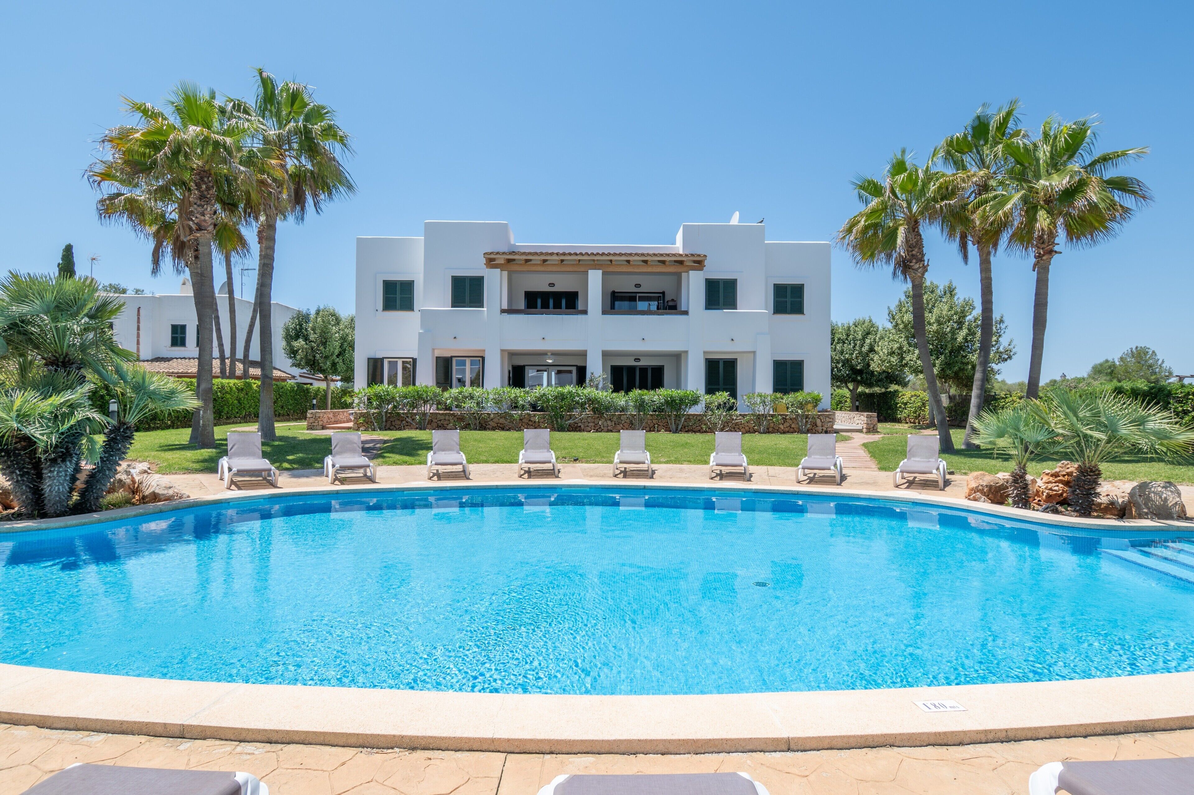 Una piscina con sillones de descanso, palmeras y un edificio blanco con balcones.
