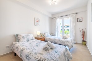 Une chambre avec deux lits, une table de chevet, une lampe, une fenêtre avec des rideaux et une plante en pot.