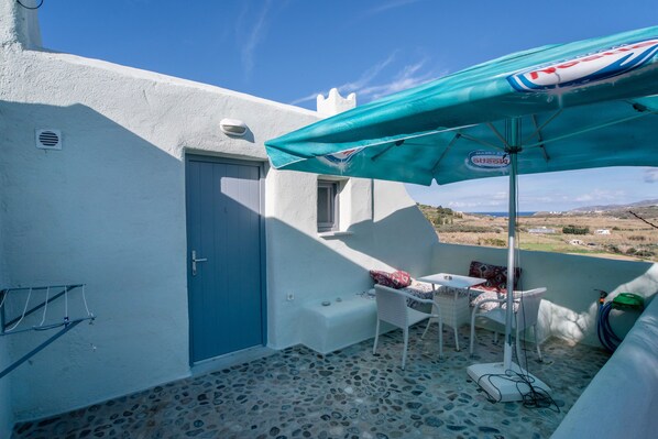 Une petite terrasse extérieure avec une porte bleue, une table surmontée d’un parasol blanc et une vue sur la campagne.