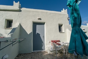 Un edificio blanco con una puerta azul, una ventana pequeña y una mesa con una silla.