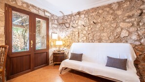 A room with a stone wall, a wooden door, a white sofa with cushions, a small table with a lamp, and a chair. - Country House 'Ses Estaques' with Mountain View, Shared Pool and Wi-Fi (Illes Balears)