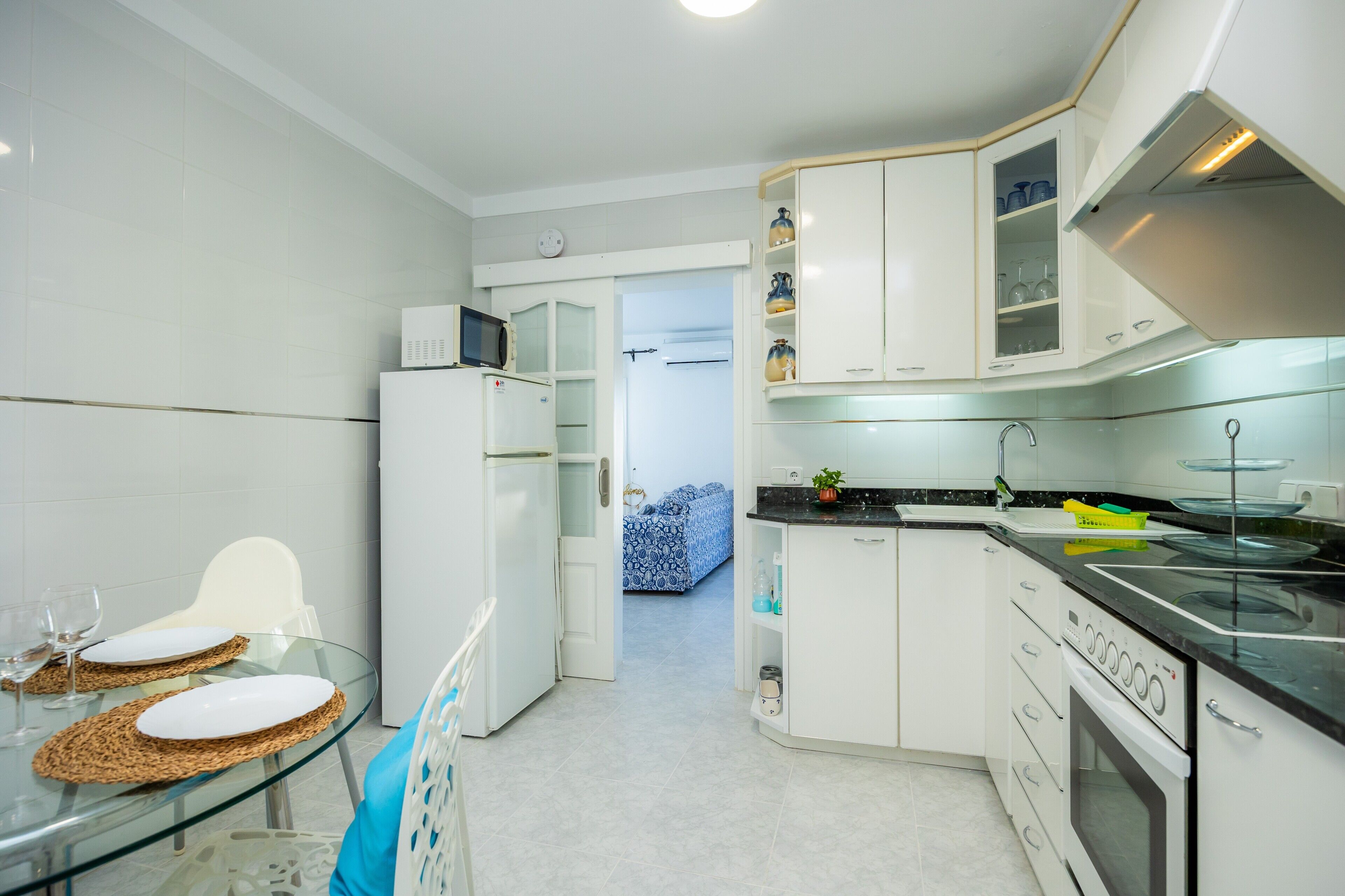 Une cuisine compacte avec des meubles blancs, un four à micro-ondes, une cuisinière et une table à manger en verre agrémentée de sets de table.
