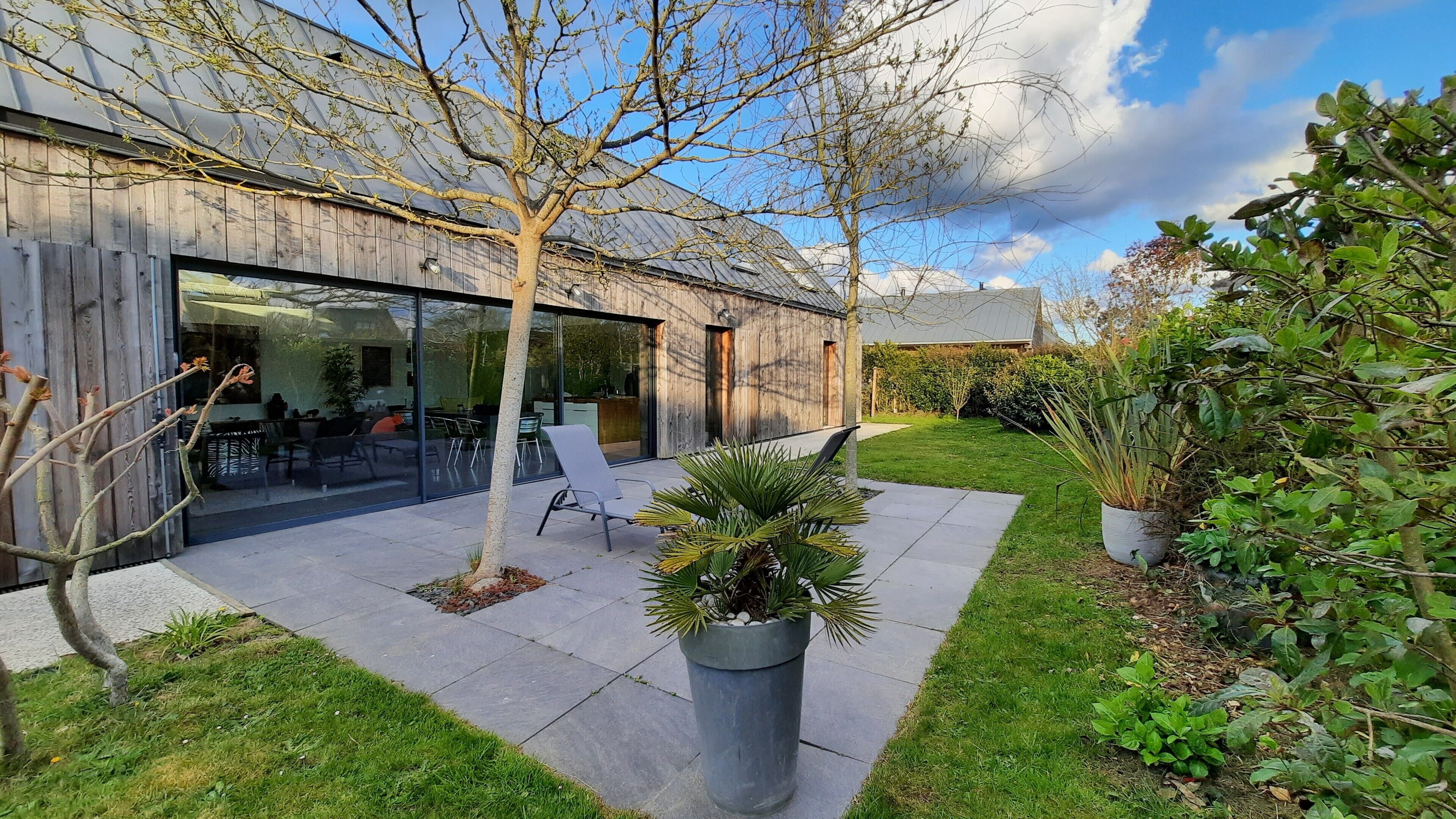 Een modern gebouw met een glazen gevel, een terras met zitplaatsen buiten, en een verzorgde tuin.