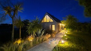 Una casa moderna con un amplio ventanal, rodeada de plantas tropicales y un sendero bien iluminado.