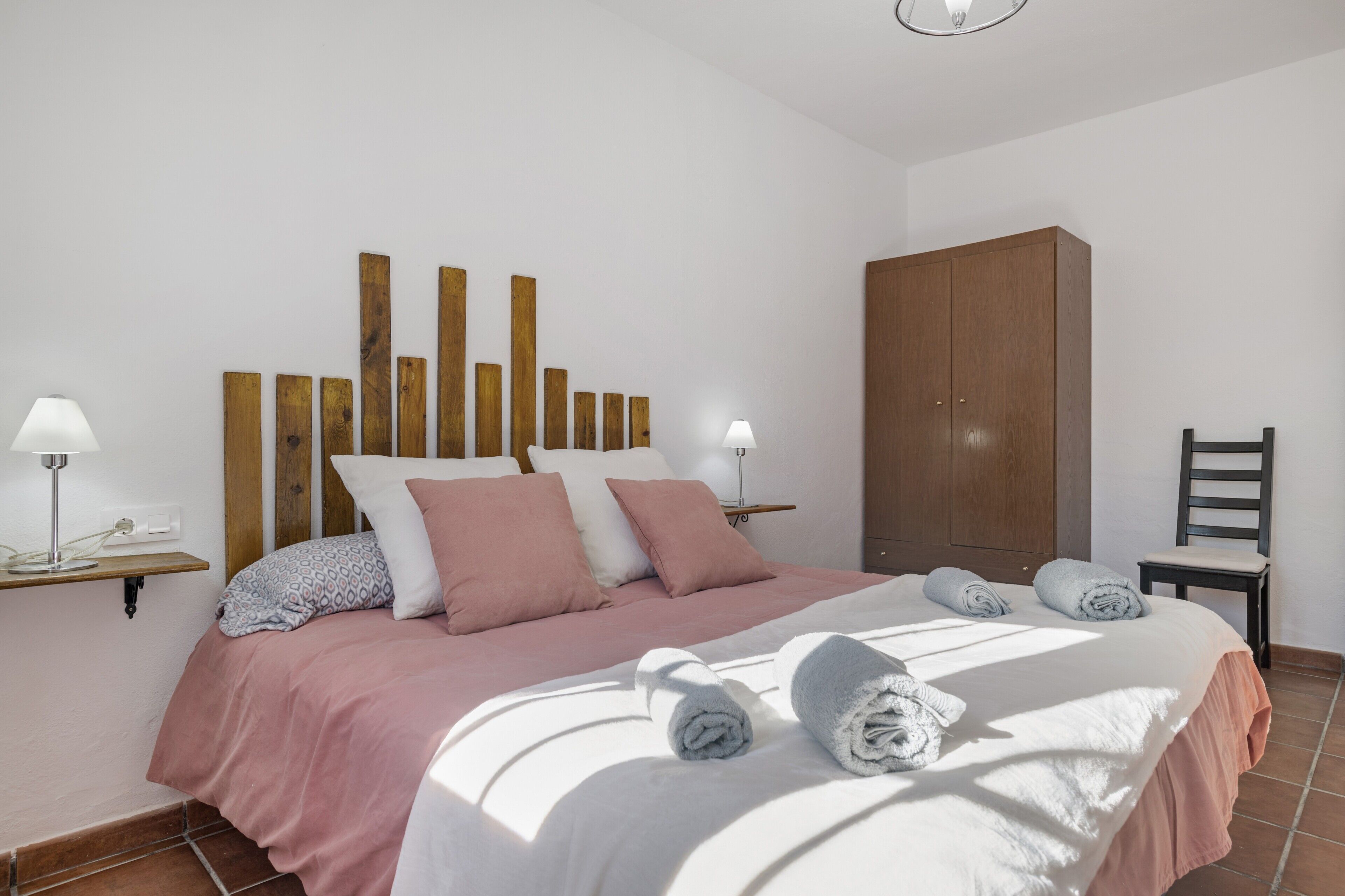 Un lit bien fait, avec des oreillers et des serviettes, une tête de lit en bois, une armoire, une chaise et une table de chevet avec une lampe.
