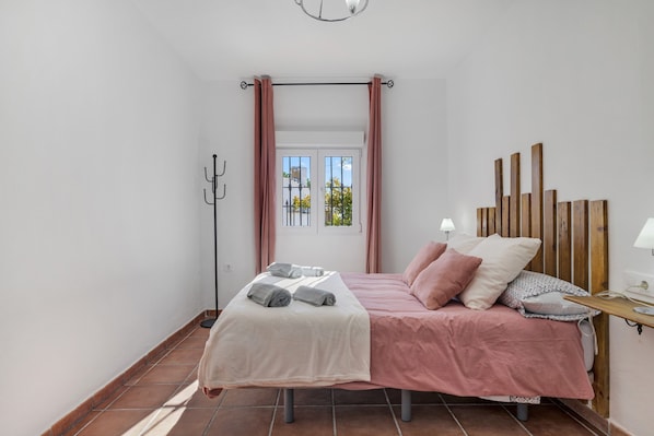 Ein Schlafzimmer mit Bett, Kissen, Nachttisch, Lampe, einem Fenster mit Vorhängen und einem Kleiderständer.