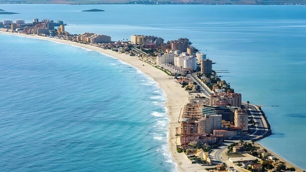 Vista aérea de uma cidade costeira com uma orla marítima, edifícios e montanhas ao fundo.
