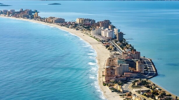 An aerial view of a coastal city with a beachfront, buildings, and mountains in the background.